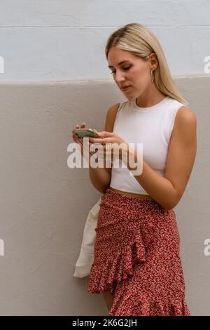 Mujer joven usando un teléfono celular en la calle de pie, apoyándose contra una pared. Foto de stock