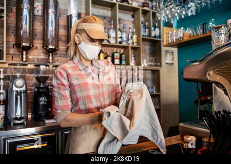 Limpieza del bar en el restaurante. Una mujer rubia adulta con uniforme de camarero se coloca detrás de la barra y limpia las copas de vino recién lavadas con un bei Foto de stock