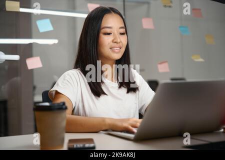 Encantadora gerente asiática mirando a la pantalla del ordenador en la habitación interior Foto de stock