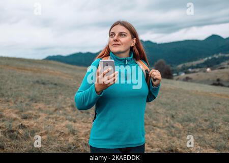 Una mujer con una mochila se coloca en la cima de una montaña con un smartphone y admira la belleza de un valle de montaña, una vista trasera. Foto de stock