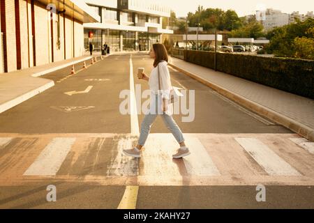 Foto de mujer joven cruzando la calle y sosteniendo una taza de café para ir. Foto de stock
