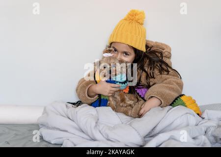 Niño vestido con abrigo beige de piel sintética cálida y sombrero de punto y bufanda sentado con un divertido perro pequeño en una cama en casa en invierno frío. Hermosa niña se calienta en manta y mira en la cámara. Foto de stock