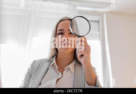 Joven mujer de negocios curiosa mirando a través de una lupa en su oficina. Empleado trabajando. Foto de stock