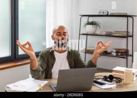 Concepto Zen. Hombre maduro tranquilo sentado en el escritorio con los ojos cerrados y meditando manteniendo las manos en gesto de mudra, oficina en casa Foto de stock