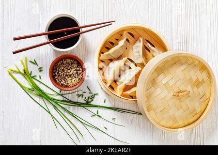 gyoza, wonton envoltorios rellenos de cerdo y col en vaporera de bambú sobre mesa de madera blanca con salsa de soja y cebolletas, vista horizontal desde arriba Foto de stock