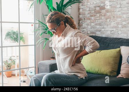 Mujer joven cansada que sufre de dolor de espalda severo sentada en el sofá en la sala de estar de la casa, mujer caucásica sentada en el sofá tocando la parte baja de la espalda sintiéndose incomodidad en casa Foto de stock