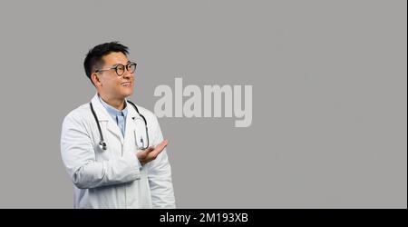 Alegre terapeuta masculino chino de mediana edad con abrigo blanco y gafas apunta al espacio libre Foto de stock
