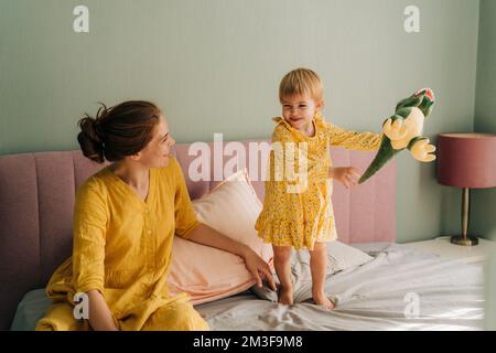 La niña pequeña riendo juega con su madre en la cama en el dormitorio. Foto de stock