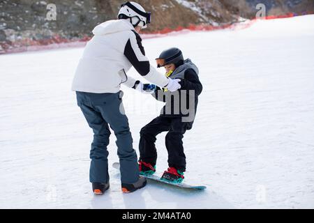Pequeño niño chino aprendiendo a hacer snowboard con su entrenador Foto de stock