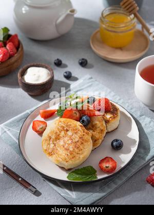 Tortitas con requesón o syrniki con arándanos frescos, fresas, miel y crema agria sobre un fondo azul con una taza de té y una tetera Foto de stock