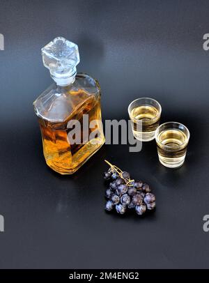 Grappa casera hecha de uvas oscuras sobre un fondo negro, una botella y dos vasos de alcohol fuerte. Primer plano. Foto de stock