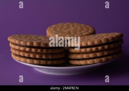 galletas redondas de foto con una capa de chocolate se encuentra apilada en un plato blanco sobre un fondo púrpura Foto de stock