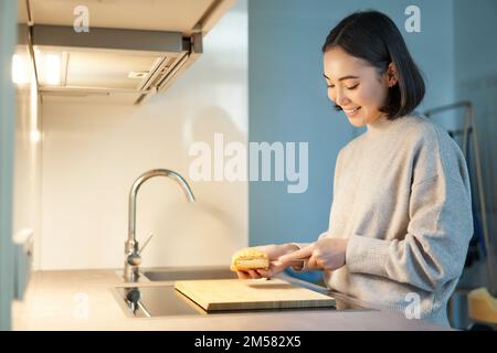 Retrato de mujer asiática joven sonriente de pie en la cocina y haciendo una bruja de arena, cocinando para sí misma