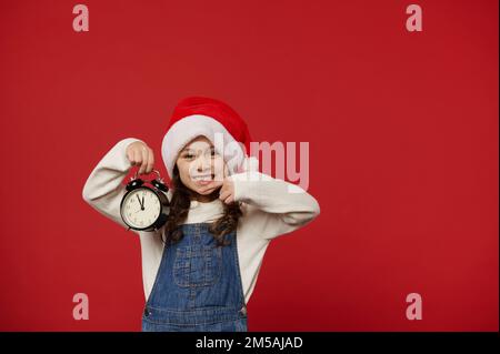 Niña alegre en sombrero de Santa, puntos en el reloj de alarma que muestra cinco a medianoche, emocionada por la próxima noche de Navidad Foto de stock