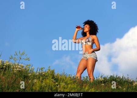 Hermosa chica africana con el pelo rizado bebiendo cerveza en un césped bajo el cielo azul Foto de stock