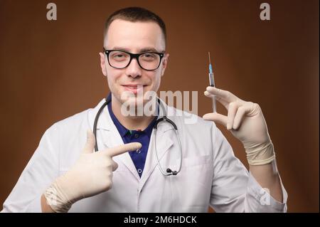 Un joven médico caucásico con una bata blanca y gafas sostiene una jeringa con una vacuna. Amable tierno joven señala un dedo A. Foto de stock