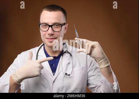 Un joven médico caucásico con una bata blanca y gafas sostiene una jeringa con una vacuna. Amable tierno joven señala un dedo A. Foto de stock
