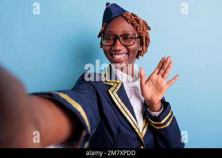 Asistente de vuelo afroamericano saludando felizmente en estudio selfie. Trabajador de la aviación que lleva gafas, mujer afro que toma el autorretrato con el teléfono móvil, smartphone. Saludo de retrato de azafata. Foto de stock