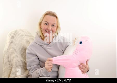 Mujer en una ropa deportiva caliente sostiene un juguete suave de tiburón rosa Ella sonríe en casa Ella abraza a un tiburón Se relaja y se duerme Descansa hablando, abrazando y besando Foto de stock