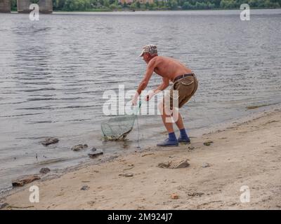 Un anciano con pantalones cortos y una gorra sosteniendo una red de aterrizaje en sus manos y sacando peces del río Dnipro en una playa urbana cerca del puente Norte en Kiev