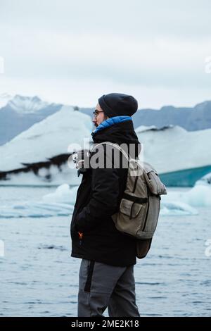 Vista lateral del fotógrafo turístico masculino en ropa de abrigo con cámara y mochila sosteniendo las manos en los bolsillos mientras está de pie en la costa pedregosa cubierta de hielo A. Foto de stock