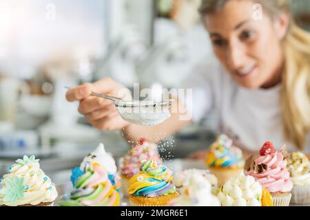 las mujeres en la panadería pastelera como confitero espolvorea el azúcar en polvo en un colador como cubierta en un muffin colorido Foto de stock