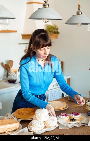 Hermosa chica en azul espolvorea el pastel con azúcar en polvo. La cocina blanca está en el fondo. Comida casera. Foto de stock