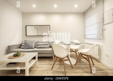 Salón-comedor de una casa amueblada con un sofá gris, una ventana con una persiana de tela y una mesa de comedor redonda Foto de stock