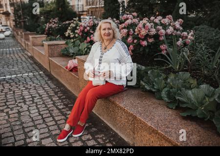 Una mujer mayor se sienta cerca de azaleas en la ciudad mientras viaja o camina. Foto de stock