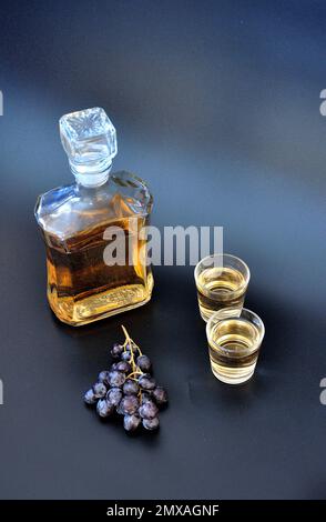 Grappa casera, alcohol fuerte en una botella de vidrio y dos vasos sobre un fondo negro, junto a un racimo de uvas oscuras. Primer plano. Foto de stock