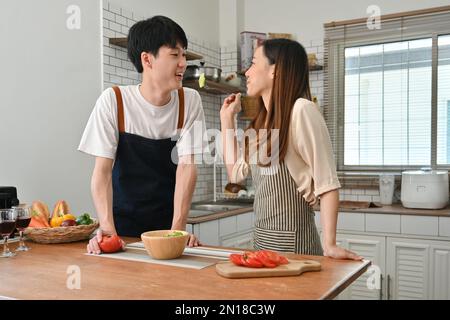 Mujer asiática sonriente que alimenta al marido, se divierten juntos en el interior de la cocina ligera. El amor, la relación, las personas y la familia
