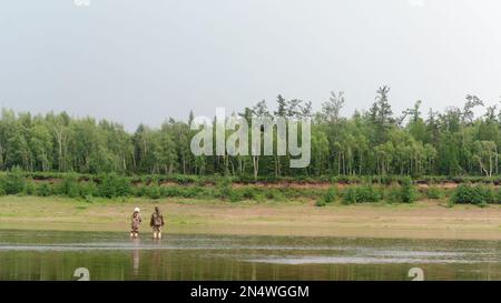 Dos Yakuts una joven y un hombre hablando, de pie en el río con una caña de pescar, enseñando a la pesca moderna girando en el fondo del bosque Foto de stock
