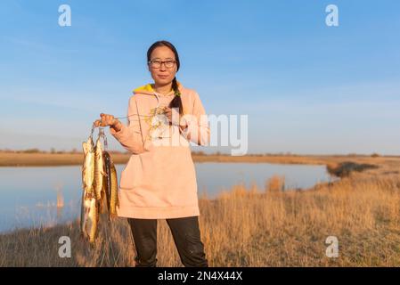 Yakut joven muchacha alegre pescador duro sosteniendo una gran cantidad de peces cogidos lucio colgando en el filete de pescado en el fondo del lago en el campo. Foto de stock
