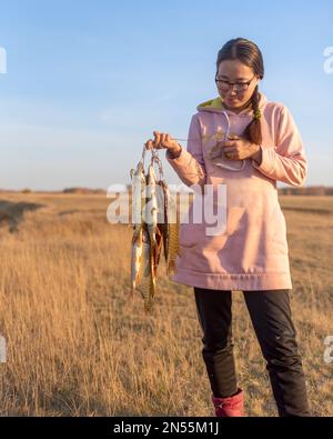 Yakut joven muchacha alegre pescador duro sostiene en sus manos una gran cantidad de pescado pavo atrapado colgando en Fish Stringer en el campo. Foto de stock