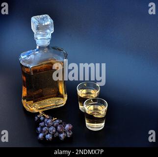 Una botella y dos vasos de grappa sobre un fondo negro, junto a un racimo de uvas oscuras maduras. Primer plano. Foto de stock