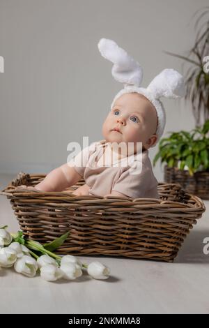 bebé en un traje blanco con orejas de conejo en la cabeza está sentado en una cesta de mimbre en el suelo con un ramo de tulipanes. Foto de alta calidad Foto de stock