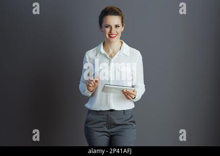 Empleado elegante feliz de la mujer de mediana edad en blusa blanca usando el APP en la PC de la tableta aislada en fondo gris. Foto de stock
