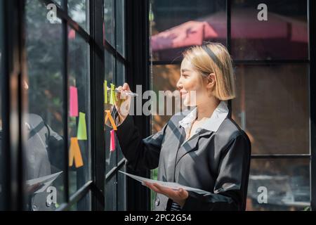 mujer asiática empresaria de pequeña empresa que pone notas adhesivas adhesivas en la pared de vidrio en la oficina durante el análisis de negocios de formulación Foto de stock