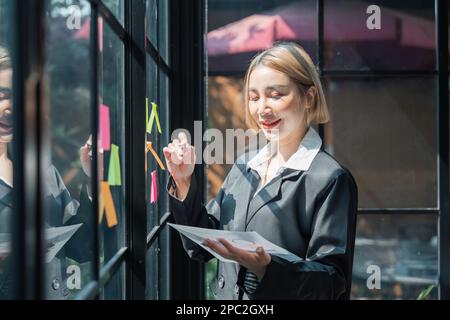 mujer asiática empresaria de pequeña empresa que pone notas adhesivas adhesivas en la pared de vidrio en la oficina durante el análisis de negocios de formulación Foto de stock