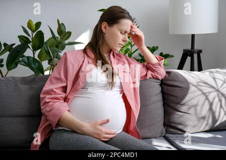 Mujer embarazada joven triste cansada que sufre migraña sentada sola en el sofá en casa. Futura mamá infeliz sosteniendo la cabeza dolorosa mientras tiene dolor de cabeza y fr Foto de stock