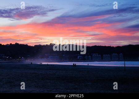 panorama de una puesta de sol en la playa donde se pueden ver los colores rojo, naranja y magenta en el cielo mientras todavía se puede ver a la gente caminando en la playa Foto de stock