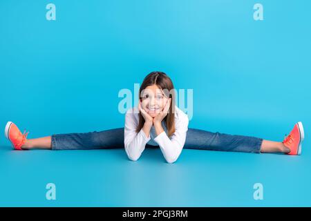 Foto de cuerpo completo de adorable colegiala vestida camisa blanca jeans sentados en brazos de hilo en mejillas aisladas sobre fondo de color azul Foto de stock