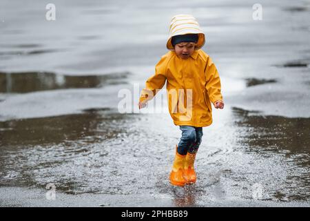 Niño con capa impermeable amarilla y botas jugando al aire libre después de  la lluvia