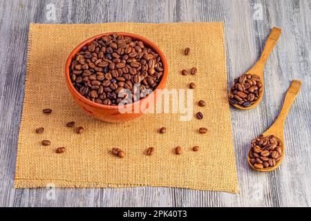 Un tazón lleno de granos de café tostados, con un puñado de granos de café crudos a su lado sobre una superficie de madera Foto de stock