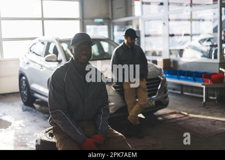 Tiro medio de un mecánico automotriz africano sonriente en su taller, su compañero de trabajo apoyado en el coche en el fondo. Foto de alta calidad Foto de stock
