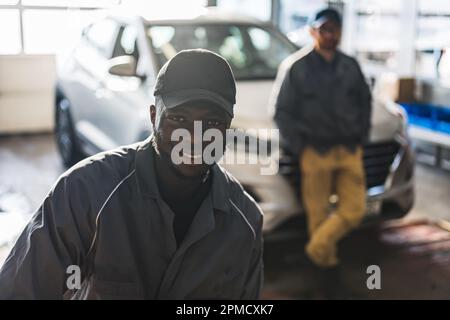 Retrato de un mecánico de automóviles africano positivo en uniforme posando después del trabajo, él está interesado en la reparación de coches, su compañero de trabajo en el fondo. Foto de alta calidad Foto de stock