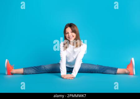 Foto de cuerpo completo de la colegiala optimista vestida con camisa blanca jeans zapatillas sentado en hilo aislado en fondo de color azul Foto de stock
