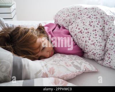 Una hermosa chica caucásica de 8 años de edad con el pelo rubio, vestida con pijama rosa, duerme en una cama con una manta esponjosa, abrazando una almohada. La luz suave del sol de la mañana fluye a través de la ventana. Foto de stock