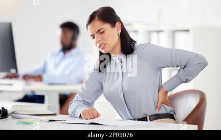 Una joven empresaria caucásica estresada que sufre de dolor de espalda baja en una oficina. Empleada femenina sintiendo tensión tensa, incomodidad y dolor en la columna vertebral con mala postura sentada y largas horas de trabajo en el escritorio Foto de stock