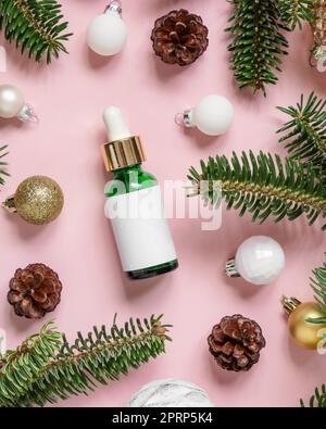 Botella cuentagotas cosmética cerca de decoraciones de Navidad, ramas de abeto y conos de pino en rosa, maqueta Foto de stock